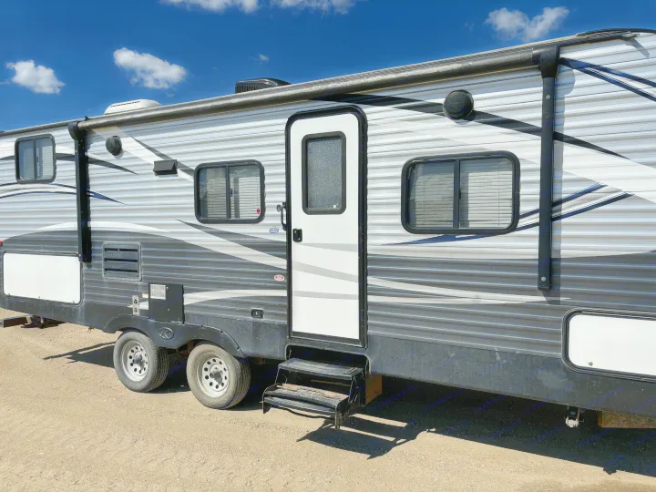 A grey and white RV rental travel trailer is parked on gravel under a blue sky. Shows door, steps, and windows.. Keystone Springdale 2017