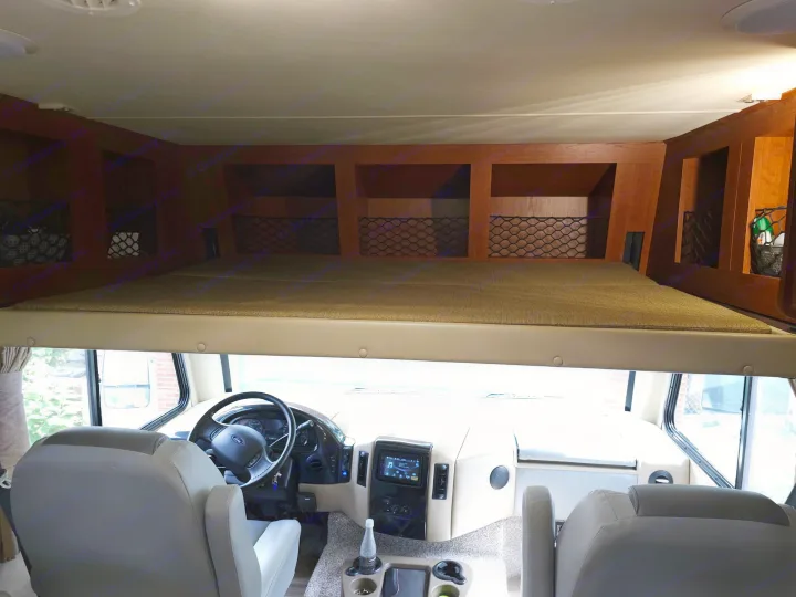 Spacious RV rental interior: view from rear showing driver's cockpit, infotainment, seats, and an overhead bunk bed with storage.. Thor 29.1 2012