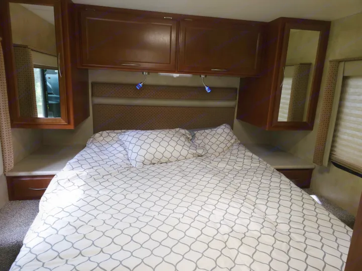 Cozy RV rental bedroom with white & grey patterned bedding, dark wood overhead cabinets, and reading lights.. Thor 29.1 2012