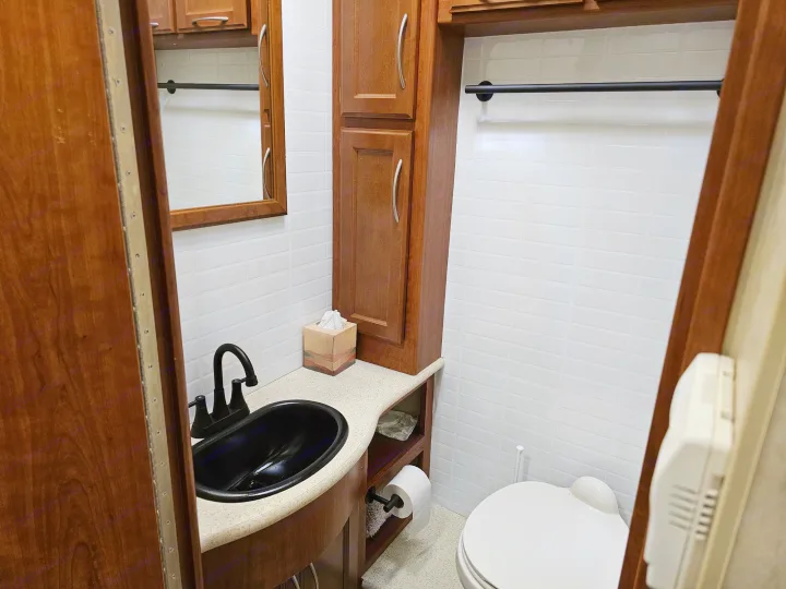 Compact RV rental bathroom with a black sink, toilet, and wooden cabinets, featuring white tiled walls.. Thor 29.1 2012