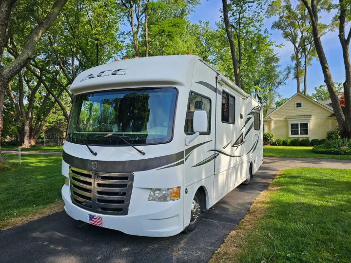 White ACE RV rental parked on a drive. Surrounded by vibrant green trees, grass, and a yellow house.. Thor 29.1 2012