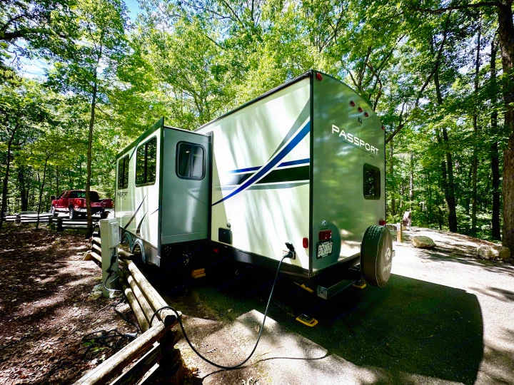 RV rental travel trailer parked in a serene wooded campsite, connected to power for a comfortable outdoor stay.. Keystone RV Passport Grand Touring 2020
