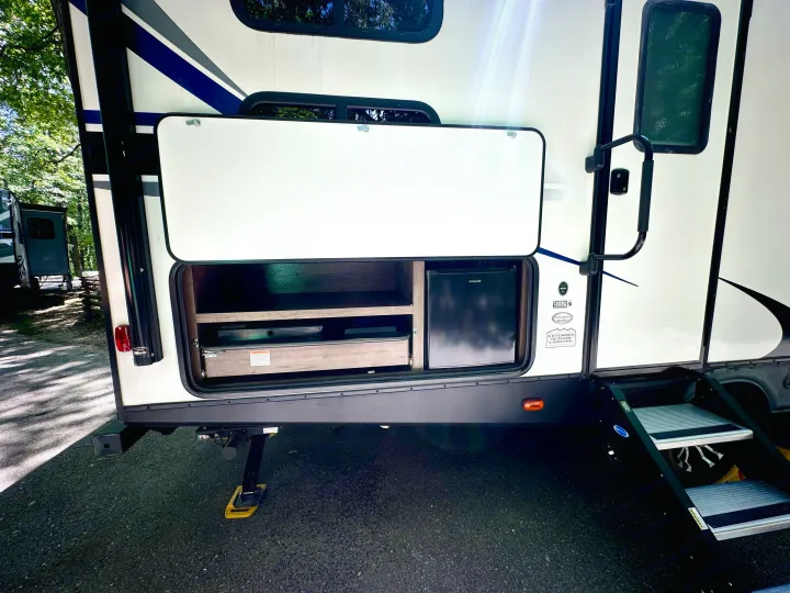 This RV rental shows an open exterior compartment with a Hotpoint mini-fridge, pull-out stove, and storage, plus entry steps.. Keystone RV Passport Grand Touring 2020