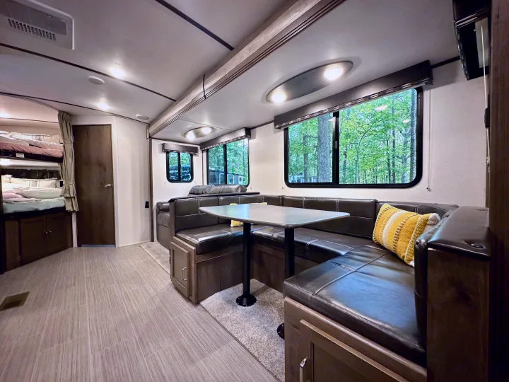Spacious rv rental interior with a dark U-shaped dinette, dining table, and windows overlooking green trees. Bunk beds seen left.. Keystone RV Passport Grand Touring 2020