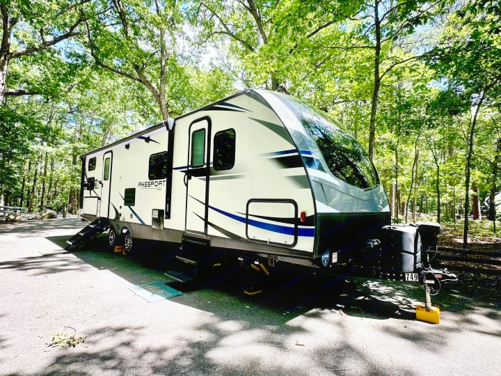 White travel trailer RV rental perfectly parked amidst lush green trees at a serene, shaded campsite.. Keystone RV Passport Grand Touring 2020