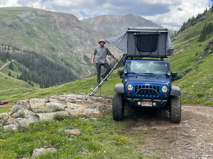 Man sets up a blue Jeep with rooftop tent on a mountain trail, perfect for an adventurous rv rental experience.. Jeep Wrangler 2010