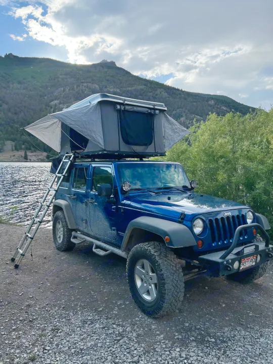 This blue Jeep with a deployed rooftop tent by a lake offers an amazing RV rental experience amidst scenic mountains.. Jeep Wrangler 2010