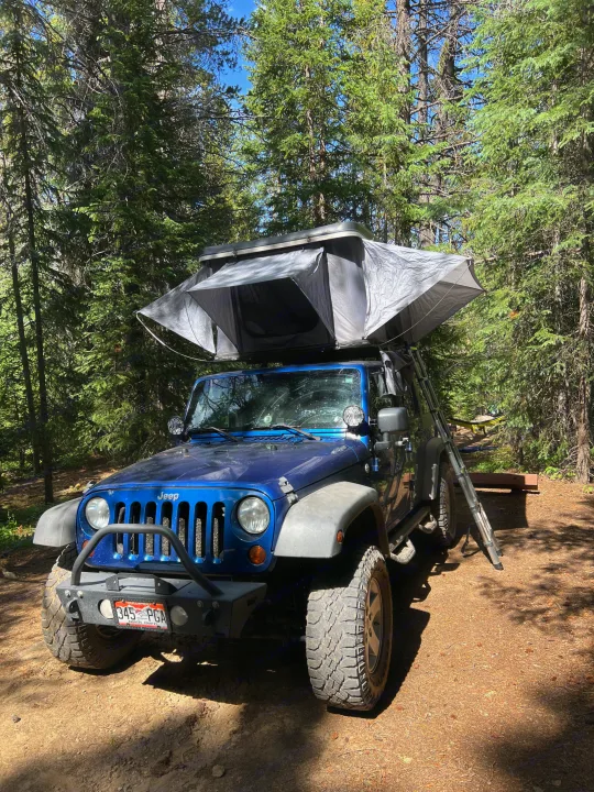 A blue Jeep Wrangler with an open rooftop tent parked in a sun-dappled forest campsite, perfect for an RV rental.. Jeep Wrangler 2010
