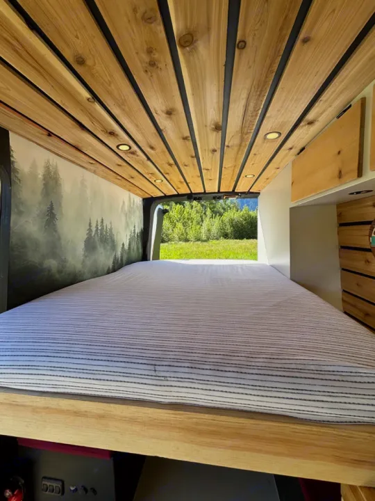 Inside a spacious RV rental: a bed with striped sheets, wood slat ceiling, forest mural, and nature view. Comfort for travel.. Nissan PRIMASTAR 2019
