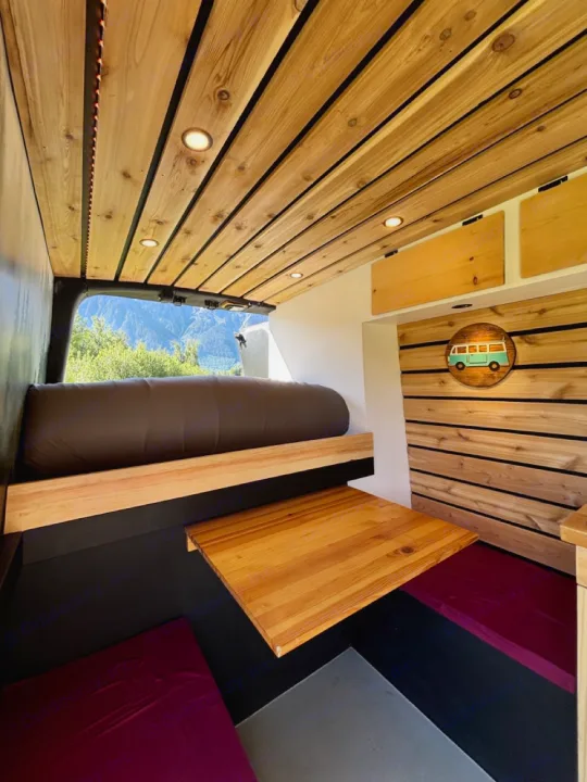 Cozy rv rental interior: wood-paneled ceiling, convertible table, comfy bed, and a mountain view from the back.. Nissan PRIMASTAR 2019