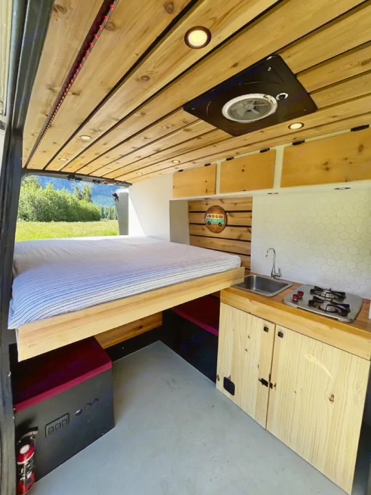 Modern RV rental conversion with wood ceiling, built-in bed, sink, stove, and overhead storage for comfortable travel.. Nissan PRIMASTAR 2019