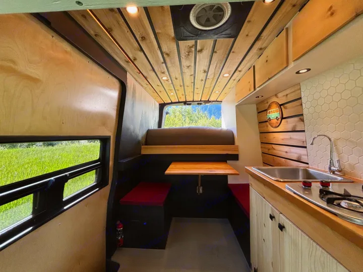Bright RV rental interior showing a wood-paneled ceiling, compact kitchen, dining area, and rear bed with scenic window.. Nissan PRIMASTAR 2019