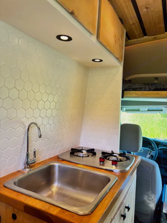 Compact RV rental kitchen with white hexagon backsplash, wood counter, sink, and two-burner stove, brightly lit. Green field seen through front.. Nissan PRIMASTAR 2019