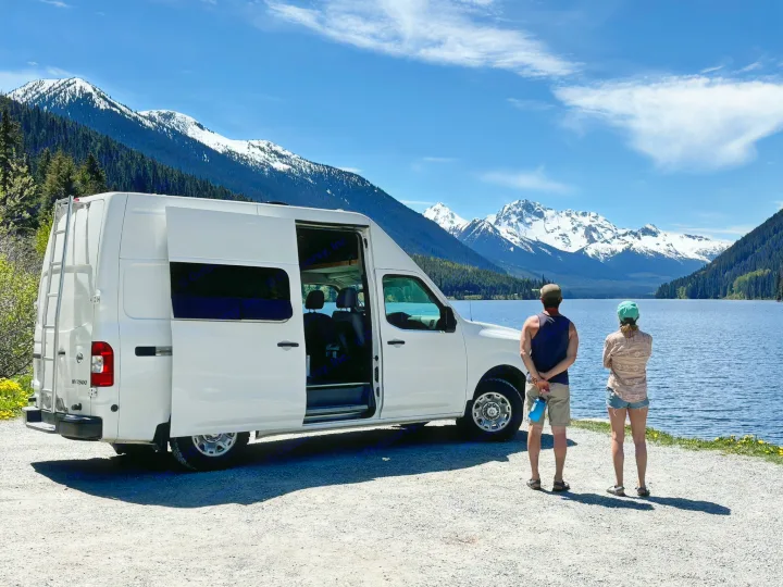 RV rental adventure: White van by a lake with snow-capped mountains. Two people admire the stunning nature view.. Nissan PRIMASTAR 2019