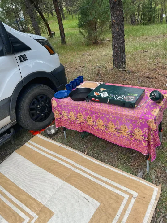 RV rental campsite with outdoor kitchen setup: propane stove, cast iron pan, camping dishes on a colorful table, and an outdoor mat.. Ford Transit 2021
