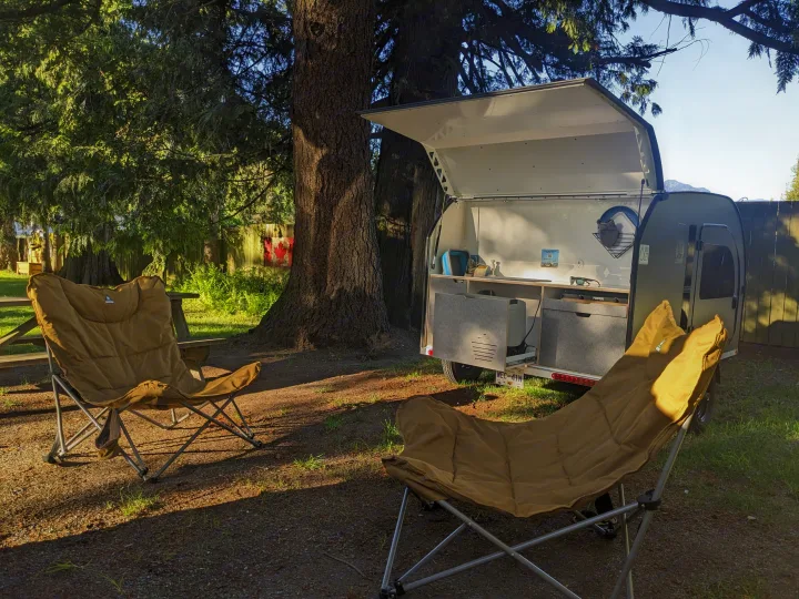 Cozy rv rental teardrop camper setup with outdoor kitchen and two chairs under large trees, perfect for camping.. DROPLET 58 2026