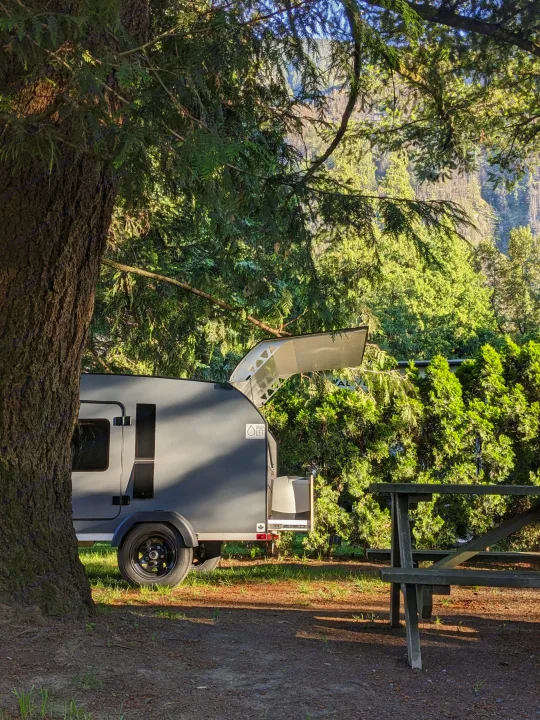 Grey teardrop RV rental parked in a sunny campsite, nestled between a large tree and a picnic table, with lush forest and mountains in the background.. DROPLET 58 2026