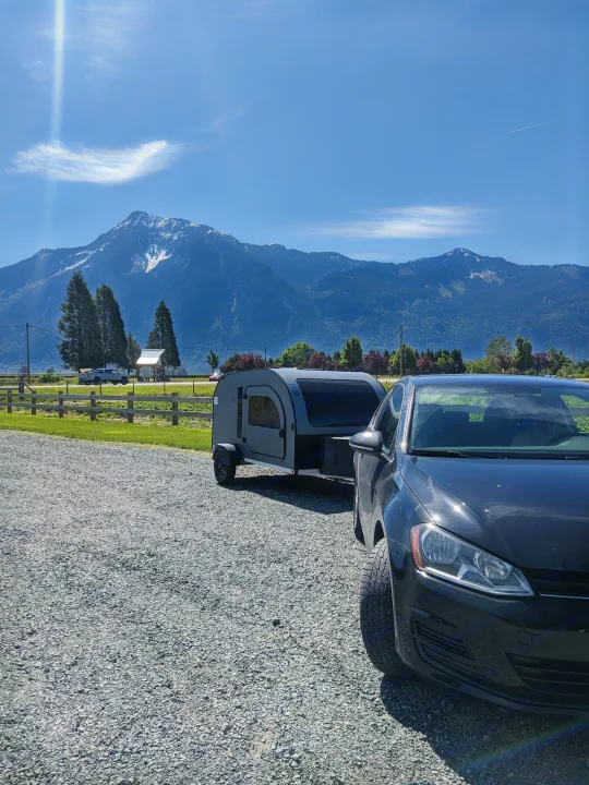 Black car towing a gray RV rental teardrop camper on a gravel lot, with majestic snow-capped mountains in the background on a sunny day.. DROPLET 58 2026