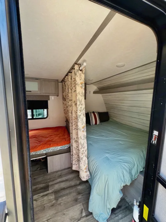 This RV rental interior features a main bedroom and a bunk area, divided by a floral privacy curtain.. Coleman Lantern 17B 2024