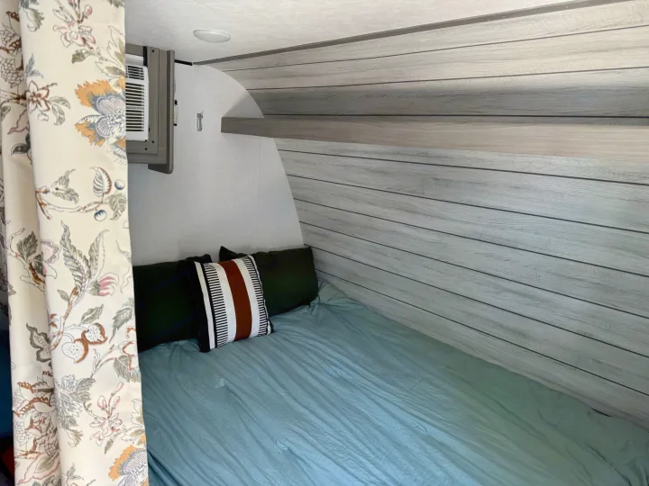 Inside this RV rental: a cozy bedroom with a bed, light blue sheets, shiplap-style walls, and an AC unit.. Coleman Lantern 17B 2024