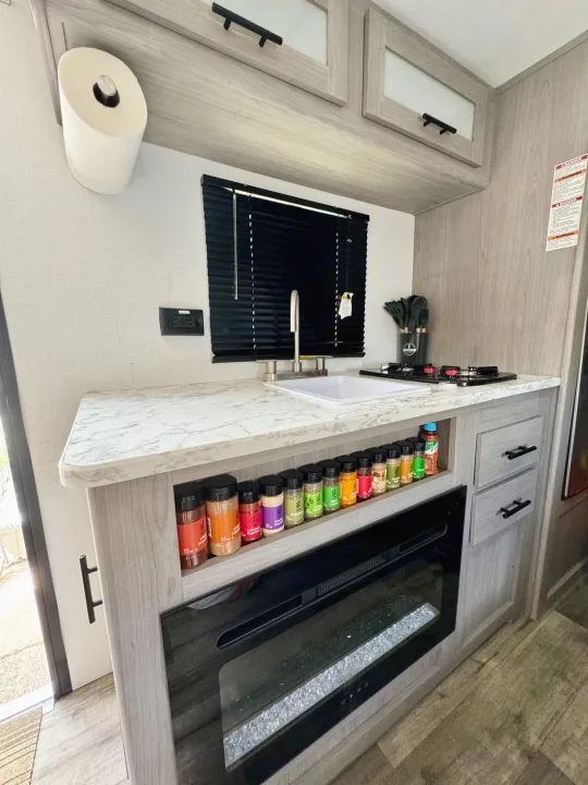 Compact RV rental kitchen: light wood cabinets, white counters, sink, stovetop, spice rack, and electric fireplace.. Coleman Lantern 17B 2024