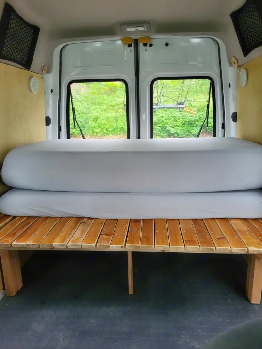 Inside an RV rental van: a cozy bed setup with wooden slats, stacked gray mattresses, and back windows.. Ford Transit 2013