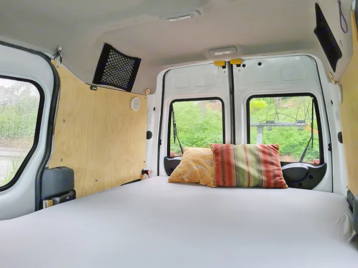 RV rental interior: A comfy bed with colorful pillows in a camper van, overlooking lush greenery through the back windows.. Ford Transit 2013