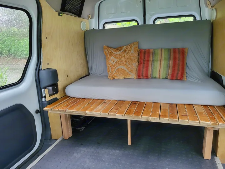 Inside a comfortable camper van, featuring a wooden slat bed/bench with a grey mattress & pillows. Ideal for an RV rental getaway, with a rainy view outside.. Ford Transit 2013