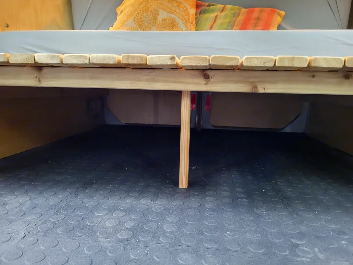 RV rental camper bed frame and storage area: A wooden slatted bed platform with a central support leg over a black rubber floor.. Ford Transit 2013