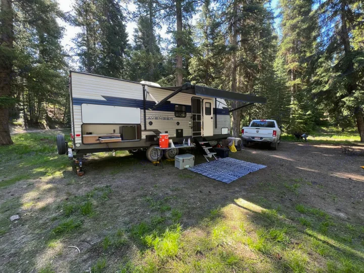 RV rental set up for camping in a forest campsite with an outdoor kitchen, truck, and an inviting mat.. Prime Time Avenger 24 BHS 2023