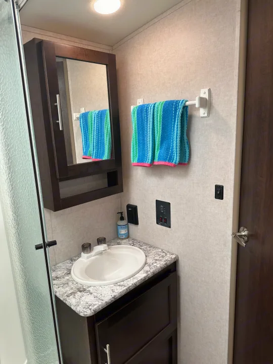 RV rental bathroom with a compact sink, mirror cabinet, and textured shower door. Features a colorful towel and control panel.. Forest River Surveyor 2018