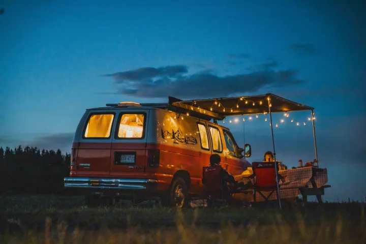 Cozy evening with an RV rental: two people relax by their van, illuminated by string lights under an awning, with a picnic table.. Ford Econoline E250supercab 2012
