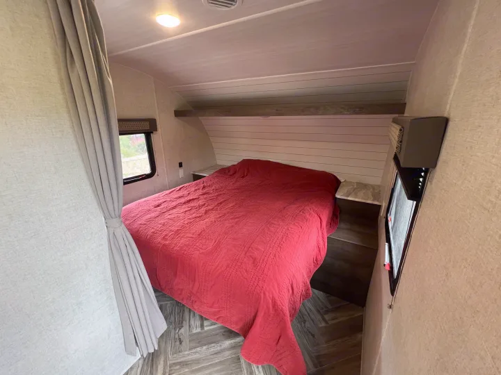 This cozy RV rental features a comfortable bedroom with a red quilted bed, built-in nightstands, and a window.. Forest River EVO 208RD 2021