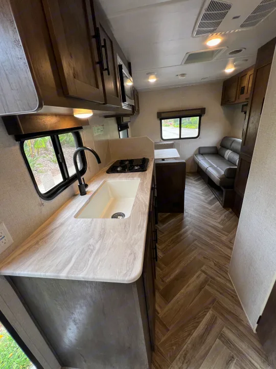 Modern RV rental interior: kitchen with sink, stovetop, and cabinets, plus a cozy living area with gray sofa and dinette.. Forest River EVO 208RD 2021