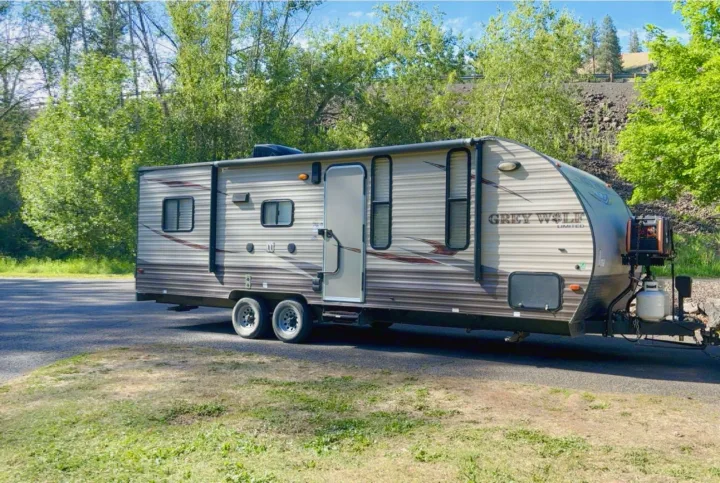 Tan Grey Wolf travel trailer RV rental, parked by green trees & hillside under blue sky. Ready for your outdoor adventure!. Forest River Cherokee Grey Wolf Toy Hauler 2016