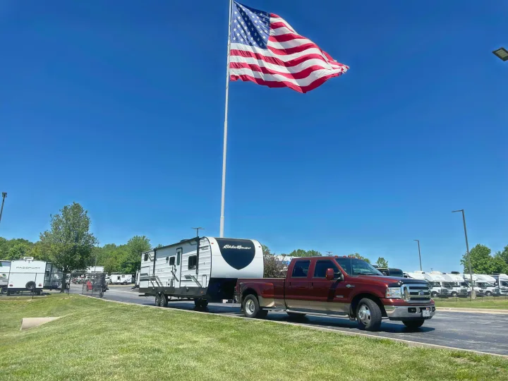 Red pickup truck towing an Eddie Bauer travel trailer at an RV dealership, with a large American flag flying high. An ideal rv rental!. Keystone RV Eddie Bauer 34bsbh 2024