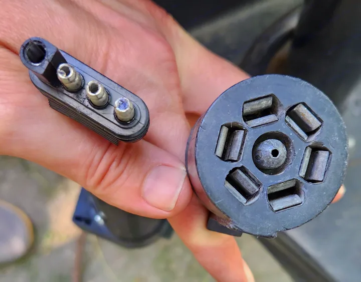 Hands holding RV electrical trailer connectors: a 7-pin round socket and a 4-pin flat plug, common for rv rental towing.. Forest River Rockwood Premier 2004