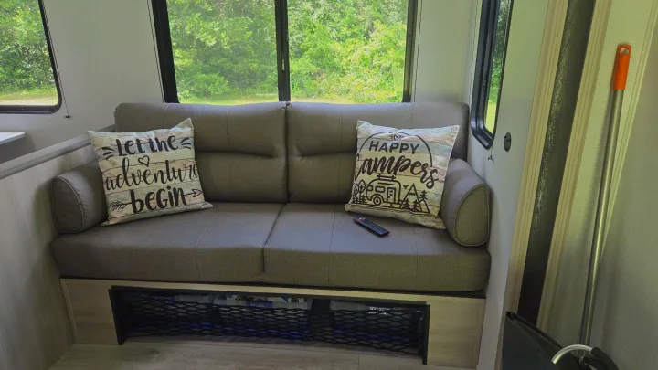 Inside an RV rental: Cozy couch with camp-themed pillows, scenic window views of trees, and under-sofa mesh storage.. East to West Silver Lake 2024