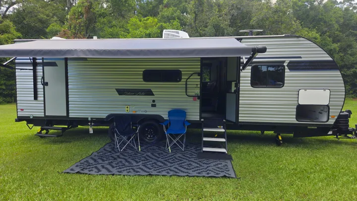 Modern rv rental travel trailer parked on green grass. Awning extended over an outdoor mat with two chairs, ready for camping.. East to West Silver Lake 2024