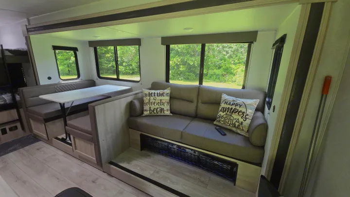 This RV rental offers a bright, inviting interior with a comfortable dinette, sofa, and large windows revealing green outdoor views.. East to West Silver Lake 2024