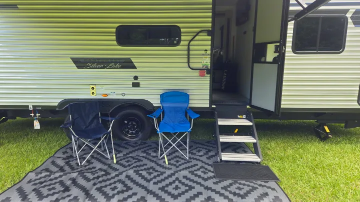Cozy RV rental setup with two camping chairs and a patterned mat on green grass, inviting outdoor relaxation.. East to West Silver Lake 2024