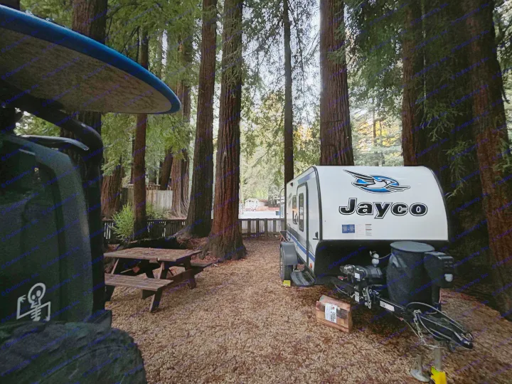 Enjoy an RV rental stay in a serene redwood forest campsite, with a picnic table, firewood, and surfboard nearby.. Jayco Hummingbird 2019