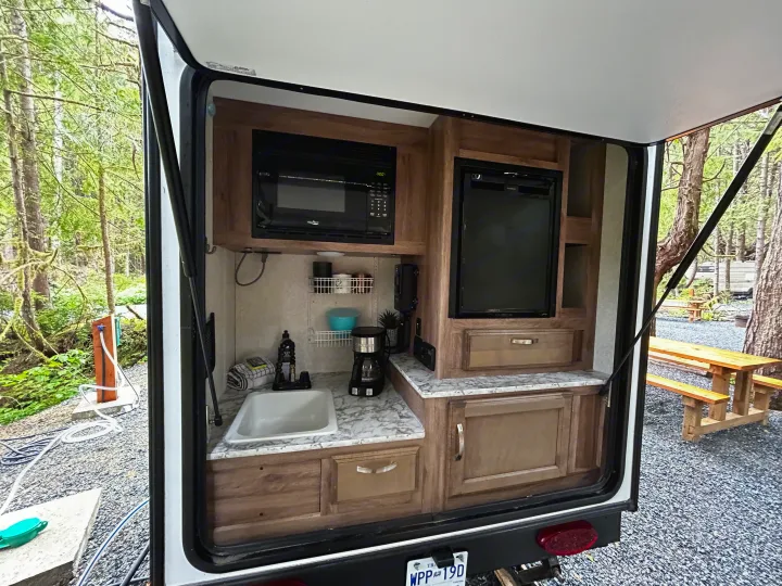 RV rental outdoor kitchen with sink, microwave, fridge, and coffee maker at a wooded campsite.. Jayco Hummingbird 2019