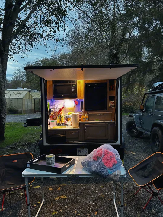 Cozy rv rental with open outdoor kitchen, camping chairs, table, and Jeep. Relaxing evening setup in nature.. Jayco Hummingbird 2019