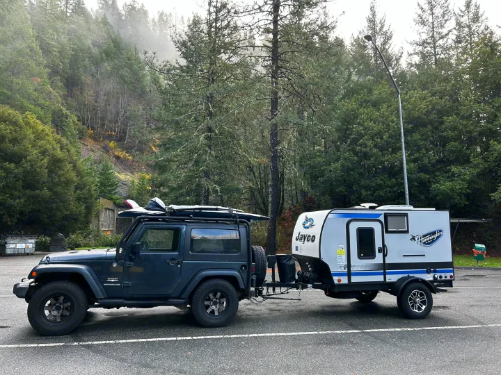 Dark gray Jeep Wrangler with surfboard tows Jayco Hummingbird RV rental trailer in a misty parking lot.. Jayco Hummingbird 2019