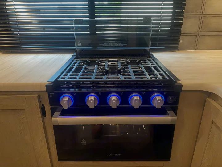 Modern RV rental kitchen features a Furrion stove/oven unit with blue LED-lit control knobs and a glass cover.. Forest River Wildwood 2022
