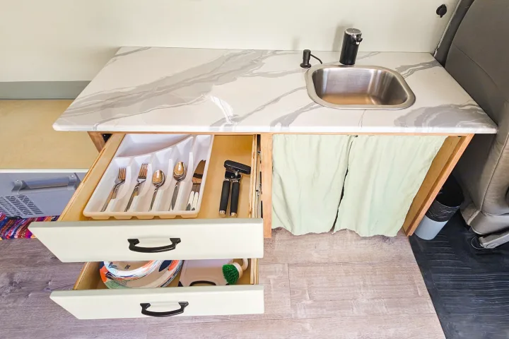 Compact RV rental kitchen with marbled counter, sink, and organized drawers for utensils and dishes.. Chevrolet Express 2500 camper 2015