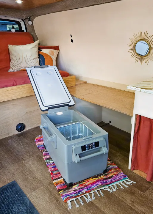 Spacious RV rental interior featuring a portable fridge on a colorful rug, wooden bed, and shelving for a comfy road trip setup.. Gmc Savanna 2003