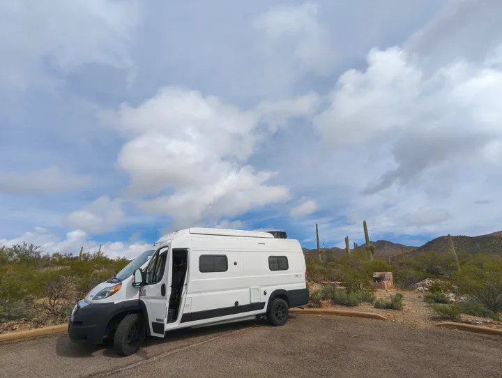White camper van (RV rental) parked in a scenic desert with saguaro cacti and distant mountains under partly cloudy skies.. Winnebago Solis 2021