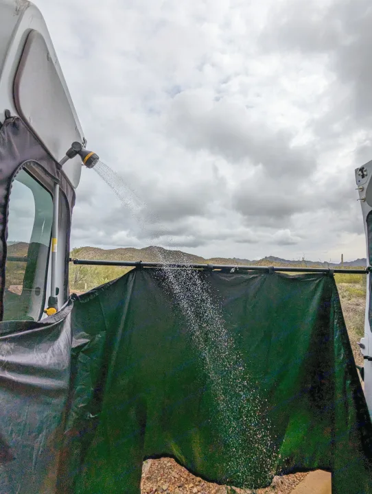 Outdoor shower in use on an RV rental van. Water sprays over a privacy curtain with a scenic desert mountain backdrop.. Winnebago Solis 2021